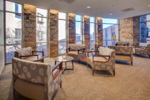 Indoor waiting area with tall windows framed by stone walls. Light, warm carpeting highlights upholstered chairs that are layout in a circle with a small coffee style table in the center.