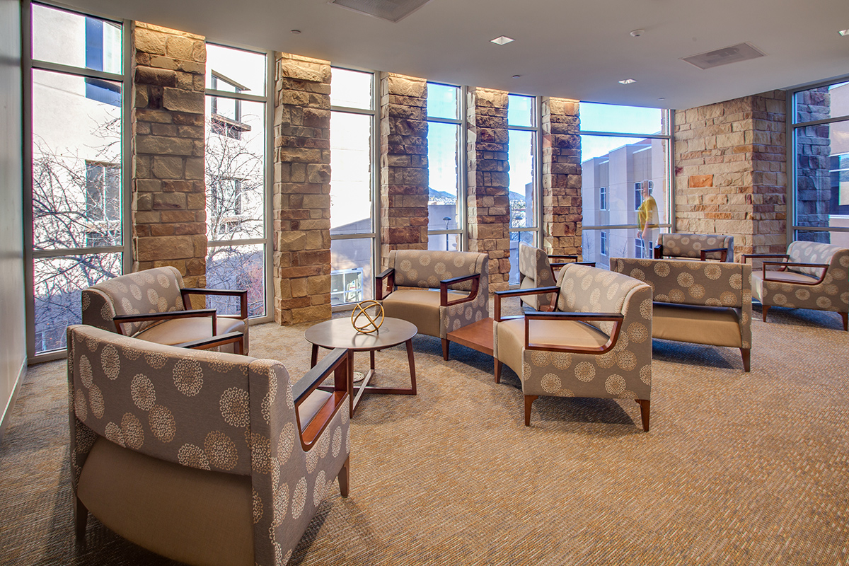 Indoor waiting area with tall windows framed by stone walls. Light, warm carpeting highlights upholstered chairs that are layout in a circle with a small coffee style table in the center.