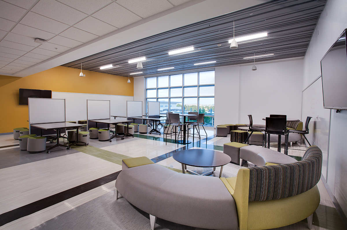 View of the common space inside of the Farmington High School. It shoes tall windows, white storage cabinets, desks and curved gray and yellow lounging spaces.